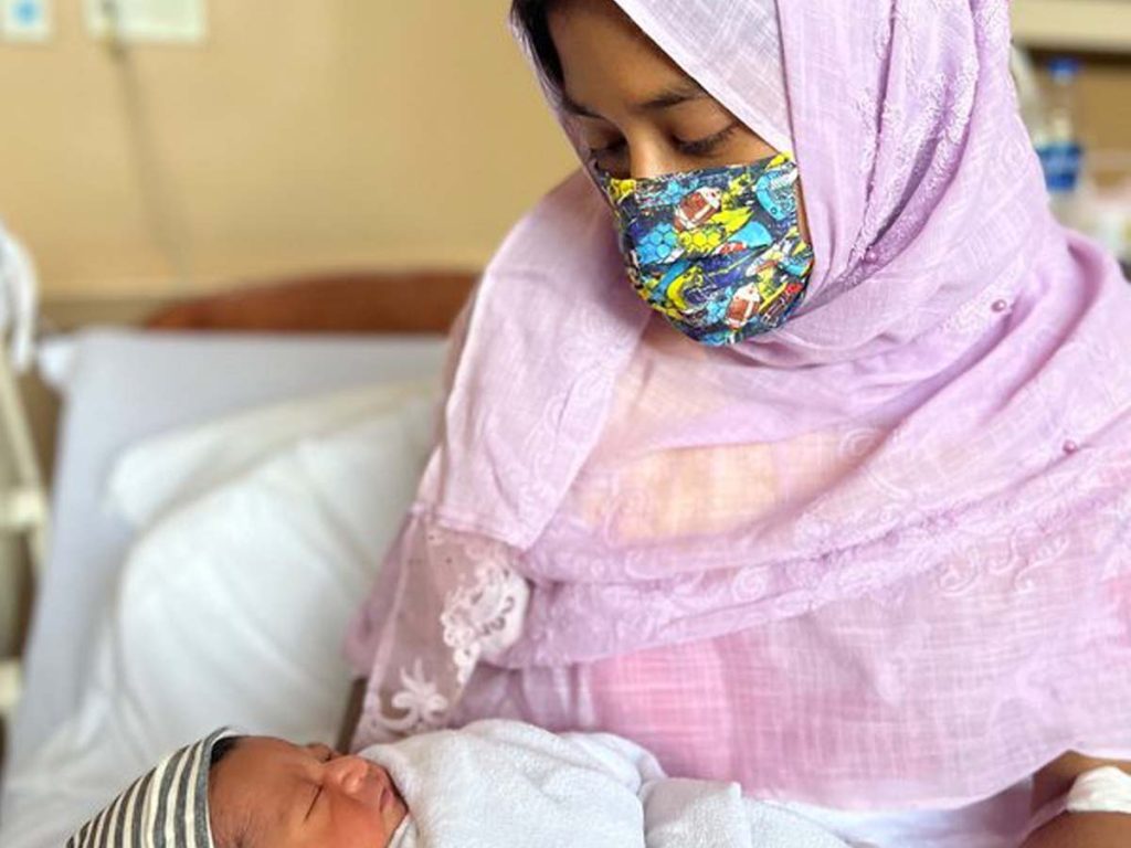 A mother sits on a hospital bed holding her newborn child.