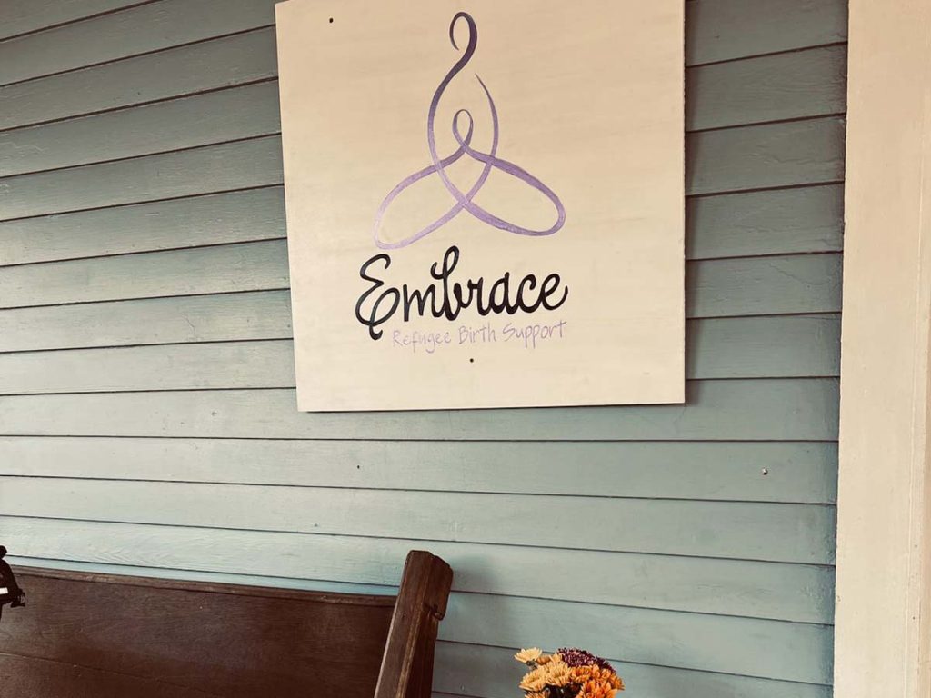 A sign with the Embrace logo hangs on a wood slat wall above a bench and a flower vase.