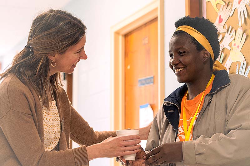 A woman hands another woman a drink in a hallway.