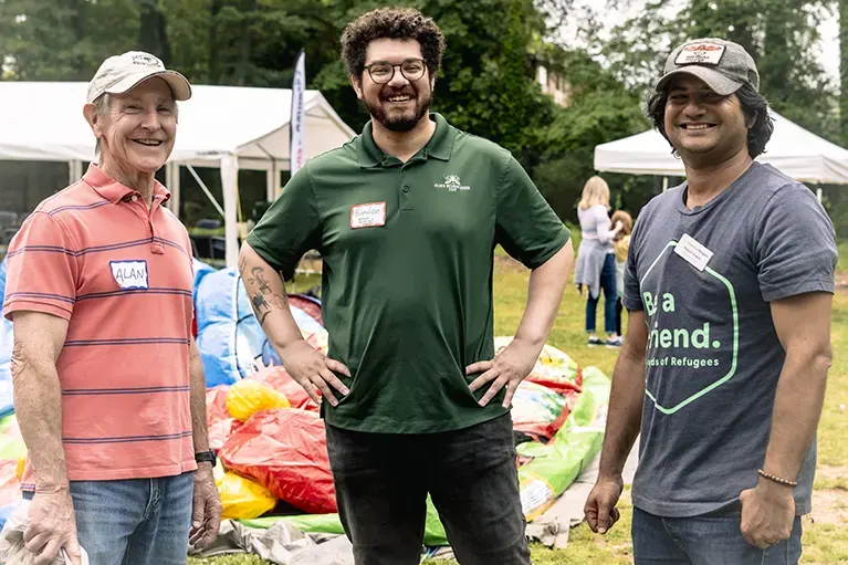 Three volunteers stand together at an outdoor Friends of Refugees event.