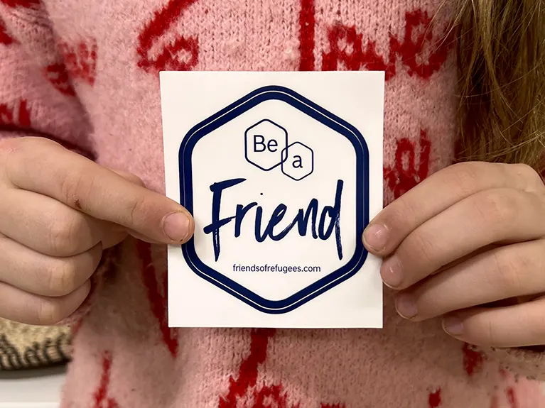 Two hands holding a sticker reading, "Be a Friend".