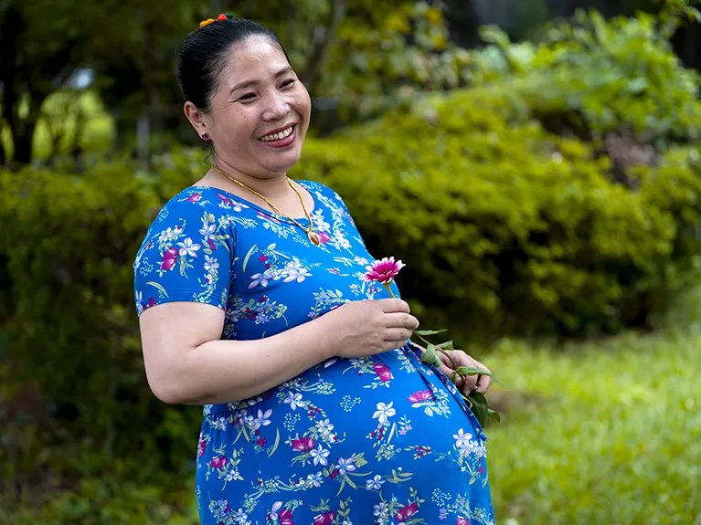 A pregnant refugee mother stands outside while holding a pink flower.