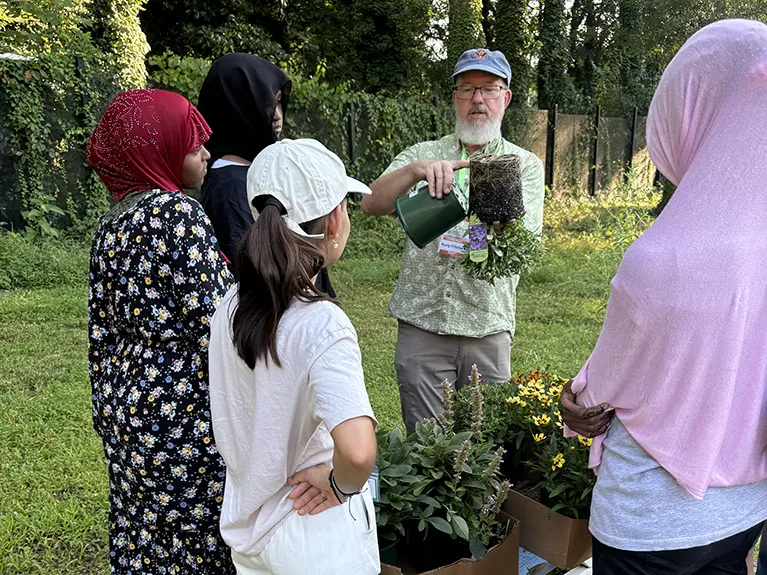Rusty Prichard holds a plant upside down and points to the roots system while instructing a group of refugees.