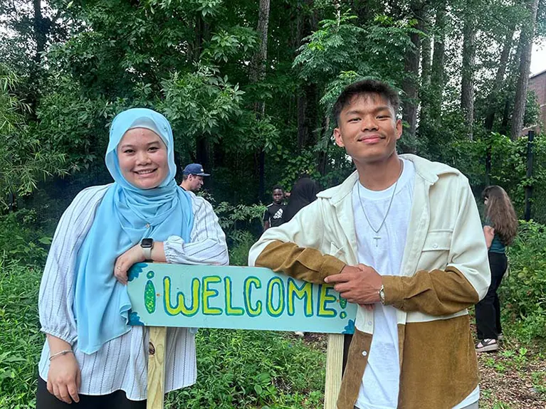 Katy and Boi pose for a photo around a hand written sign that reads, "Welcome" at the Jolly Avenue Garden.