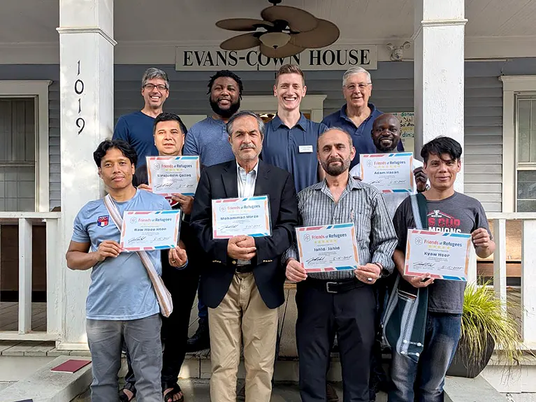 A group of volunteers and students stand on a house porch displaying their class completion certificates.