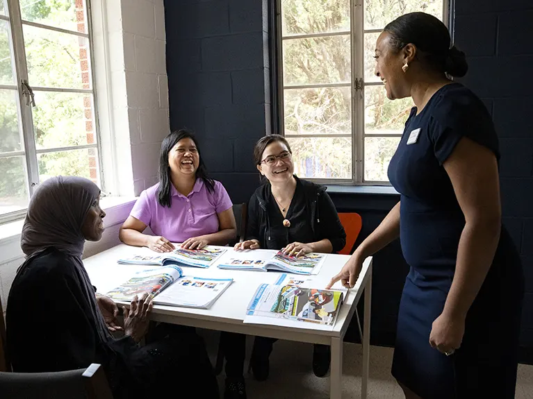 A Friends of Refugee staff member leads three women sitting around a table with workbooks in a literacy class.