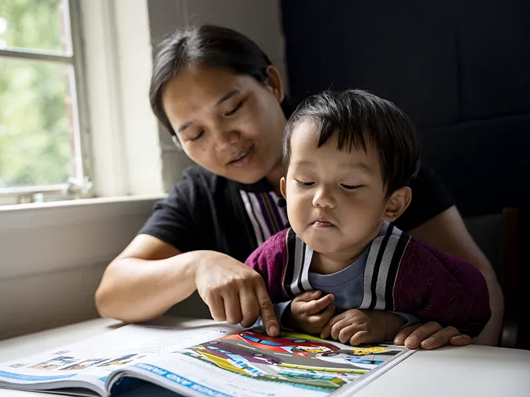 A refugee mother and her young child sit at a table reading a book together.