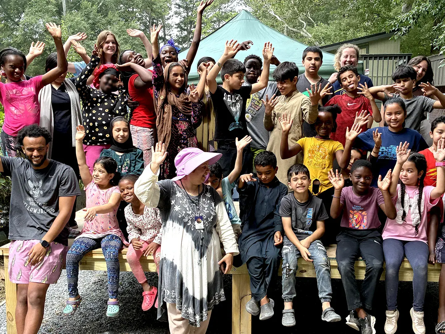 A large group of kids and adult volunteers at a Friends of Refugees youth program event wave at the camera.