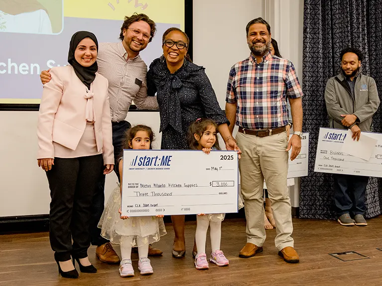 A Start:ME graduate poses with his family while his two small daughters hold a large sign that looks like a bank check.