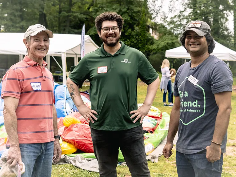 Three volunteers stand together at an outdoor Friends of Refugees event.