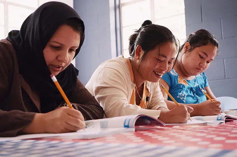 Three refugee women write in workbooks at a table while attending a literacy class.