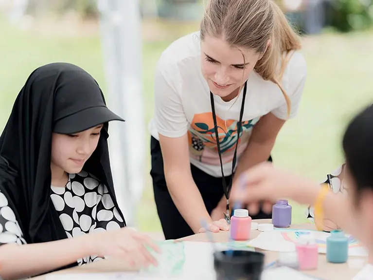 A volunteer check in on a refugee student working on an art project at a table at a Refugee Youth outdoor program.