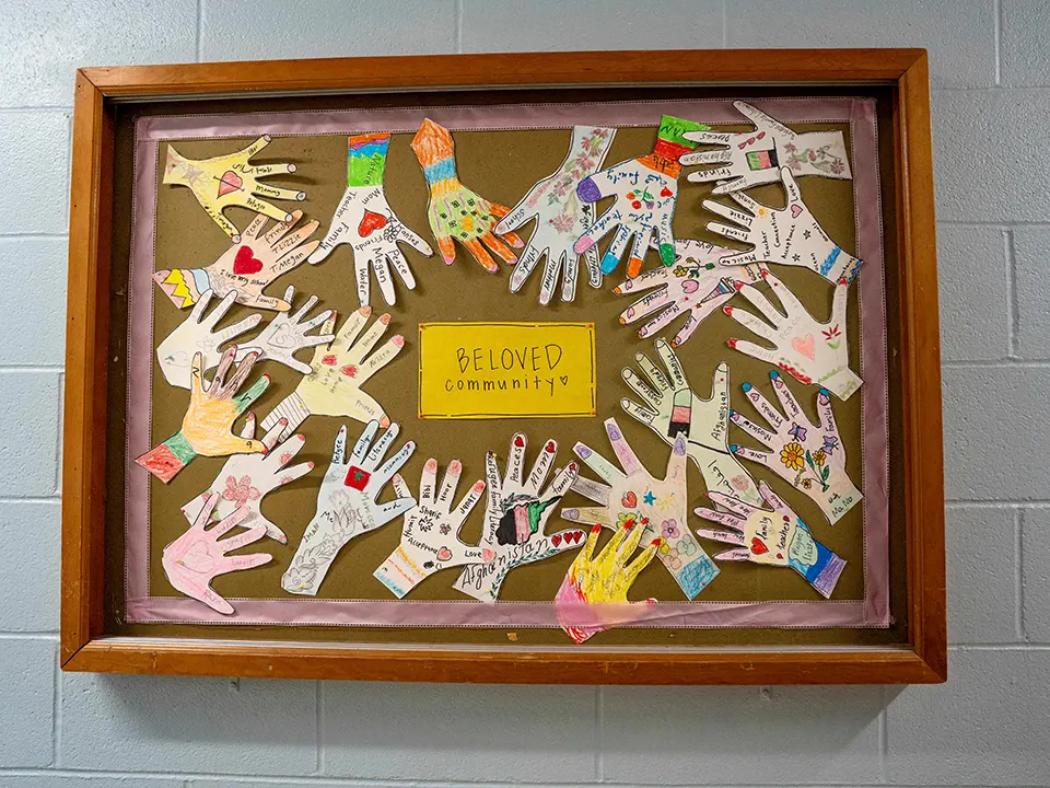 Artwork hanging on a wall with cut out paper hands and a yellow rectangle in the center that reads, "Beloved Community".