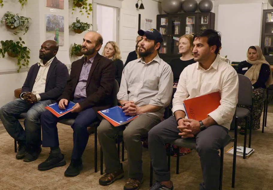 A small group of adults sit in a classroom listening to a speaker.